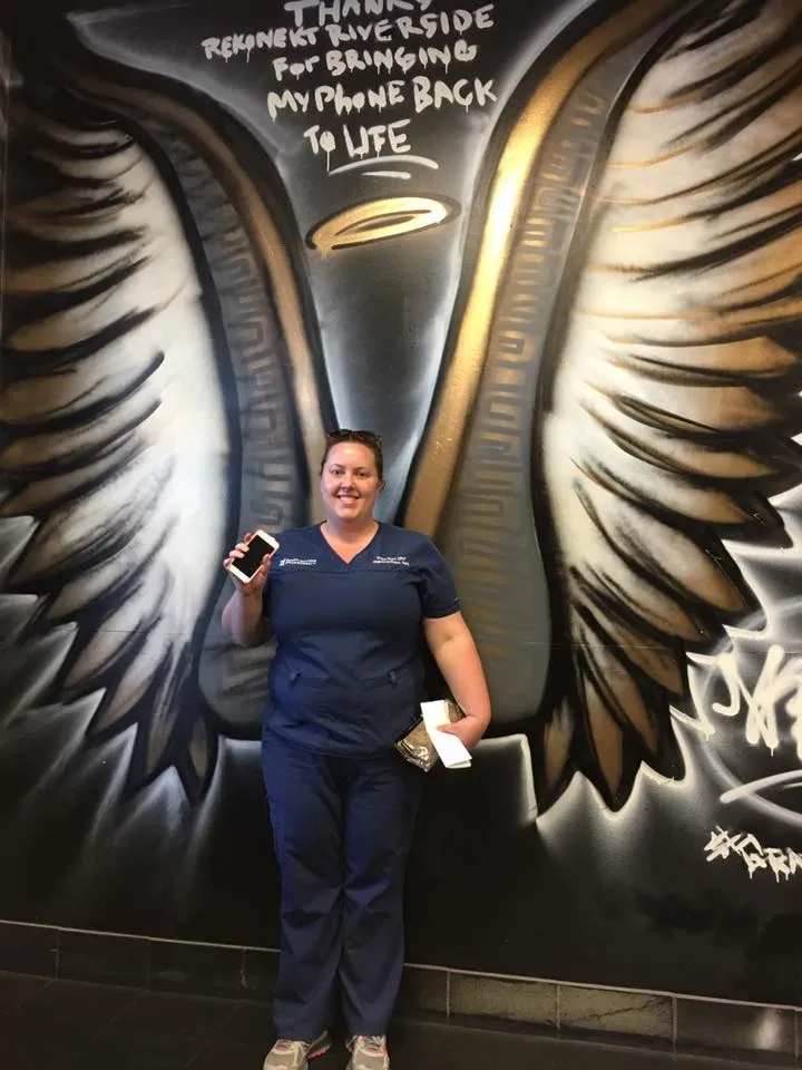 Smiling woman in blue scrubs holds a phone, standing in front of a mural of large angel wings and graffiti text.