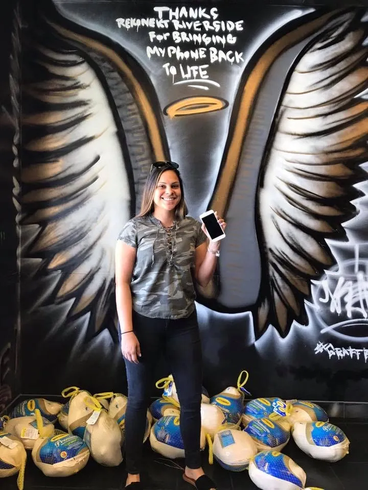 Woman stands smiling, holding a phone in front of a mural with wings and text; bags sit on the floor around her.