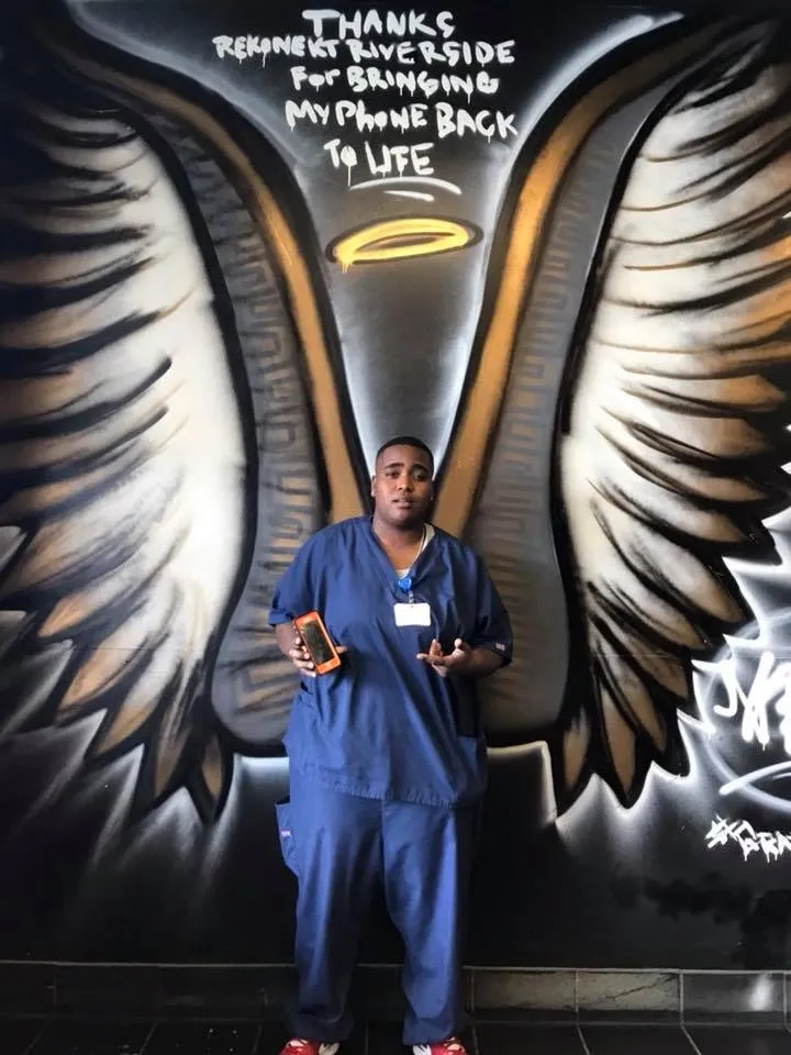 Man in blue scrubs holds a phone, standing in front of a mural with angel wings and a thank-you message above his head.