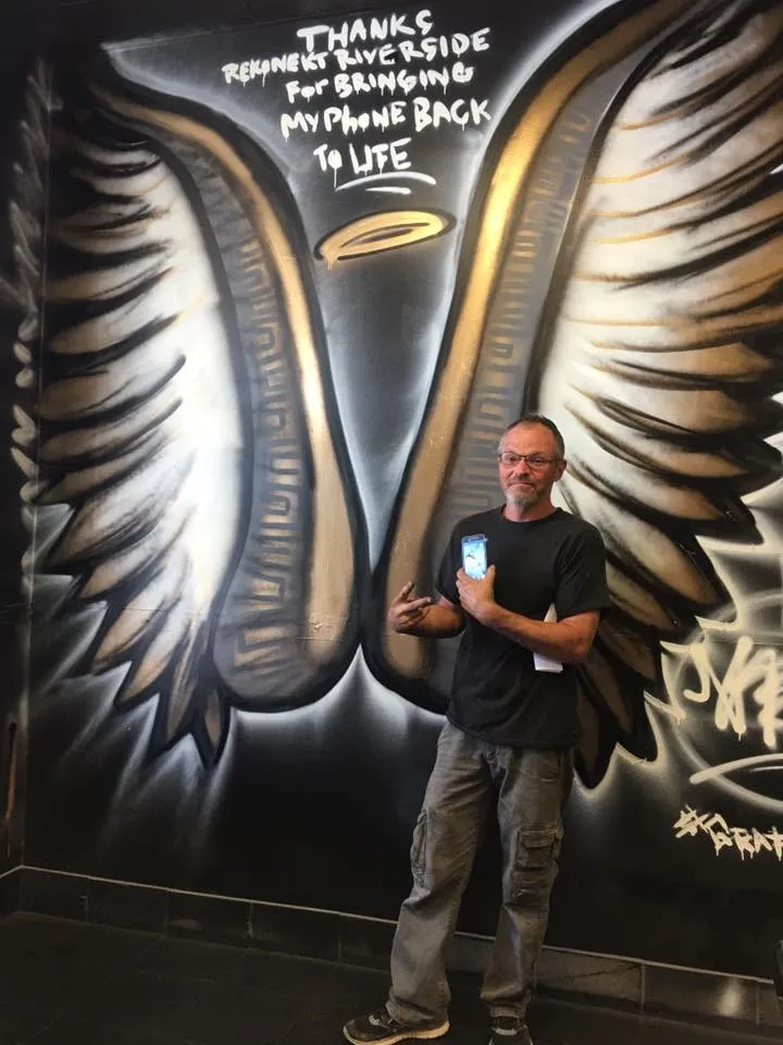 Man stands in front of a mural with wings and a halo, holding a phone and smiling at the camera.