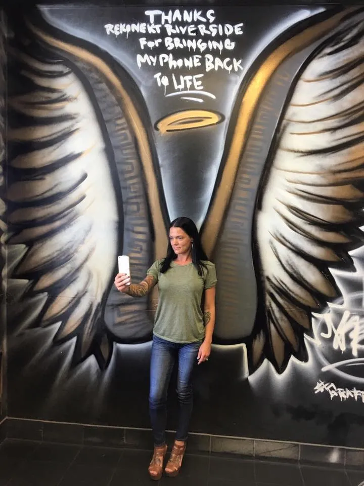 Woman taking a selfie in front of a mural with angel wings and a halo painted on a wall with a thank-you message.
