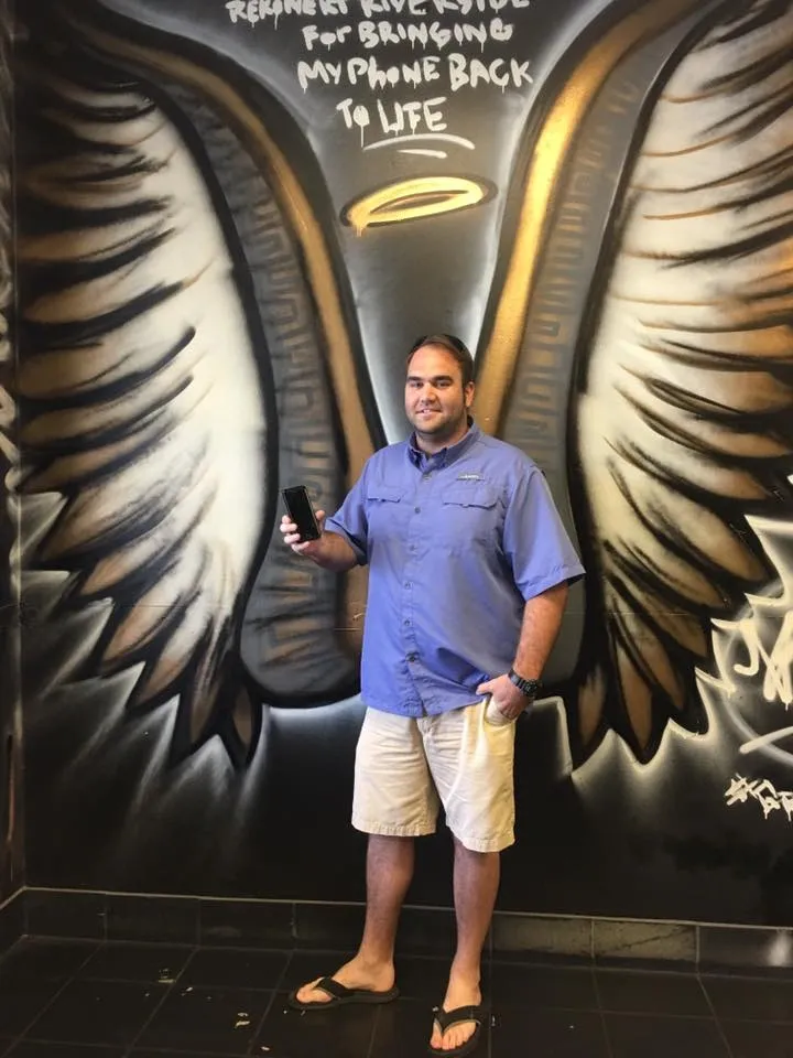 Man in shorts and flip-flops poses holding a phone in front of a mural of angel wings and text on a wall.