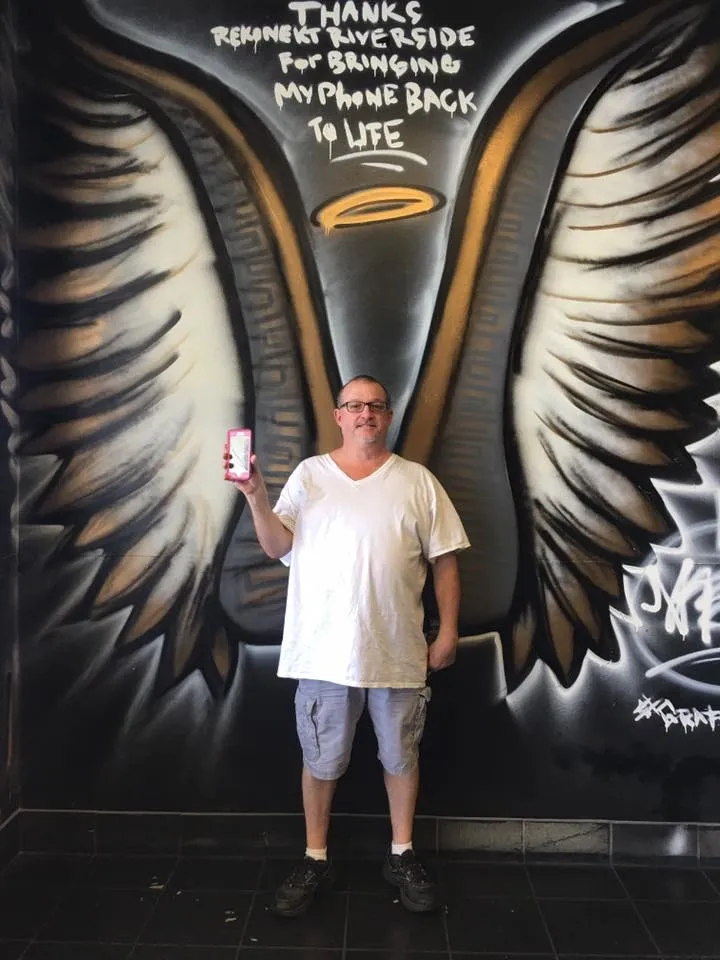 Man in white shirt stands in front of wing mural, holding up a phone, with graffiti text above him on the wall.