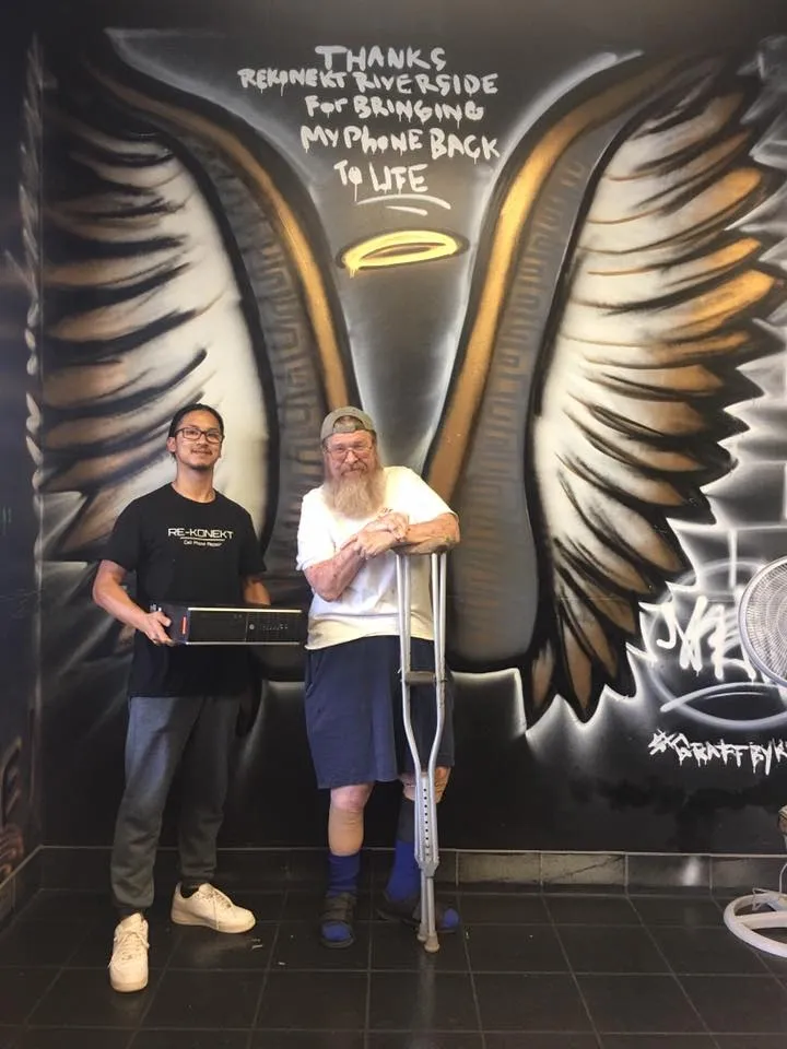 Two men stand in front of a mural with wings; one holds a box, the other uses crutches and smiles.