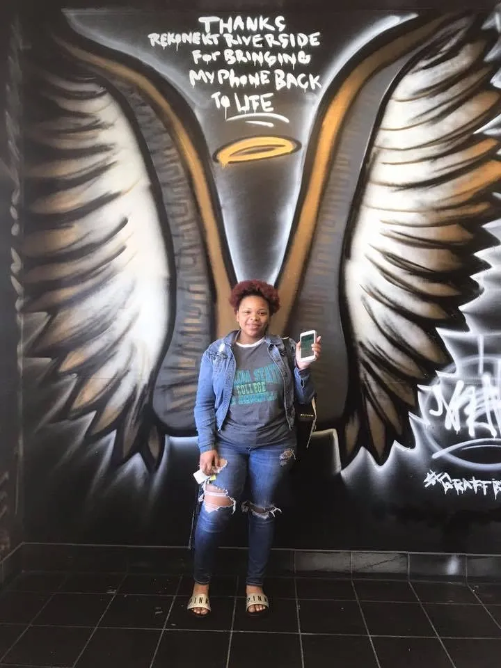 Person stands in front of angel wings mural, holding a phone and smiling, with a gratitude message painted above.