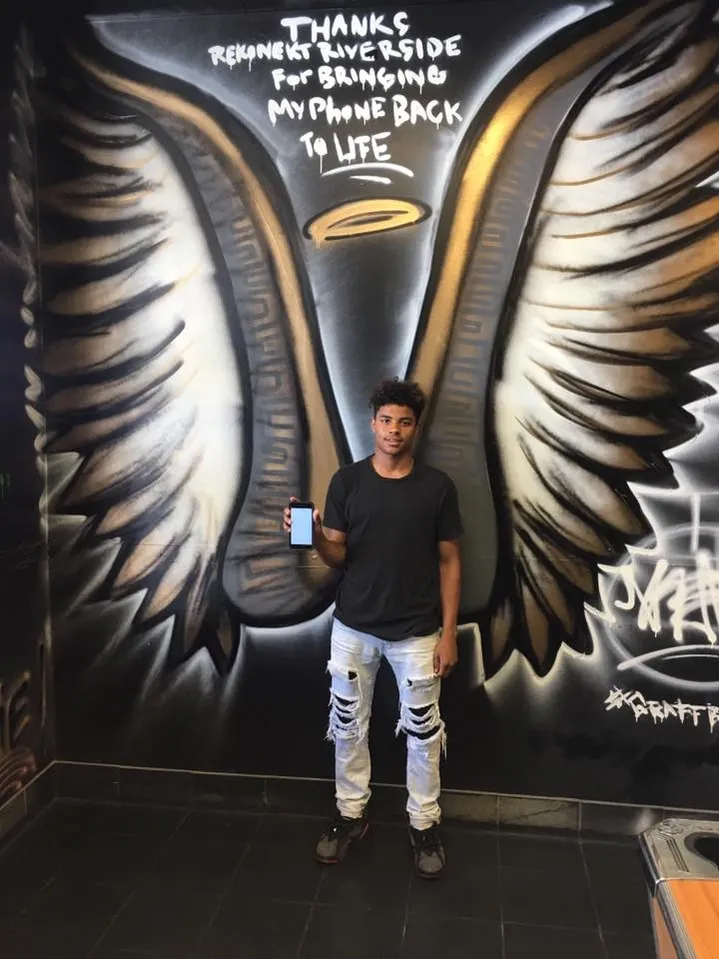 A man holds a phone in front of a mural with large wings and text thanking someone for fixing his phone.